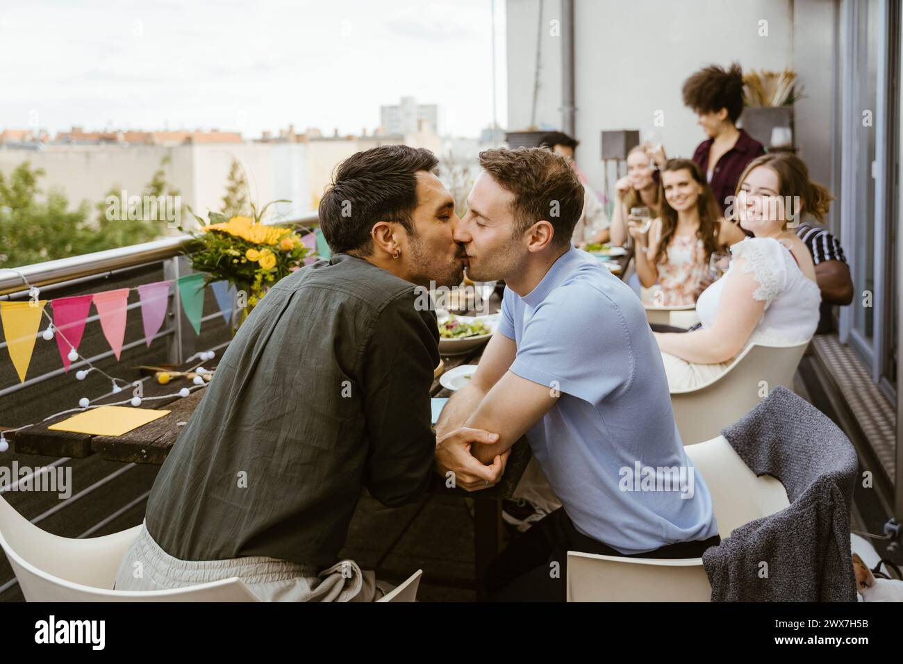 Liebevolles schwules Paar küsst sich, während es mit Freunden auf einer Dinnerparty auf dem Balkon sitzt Stockfoto