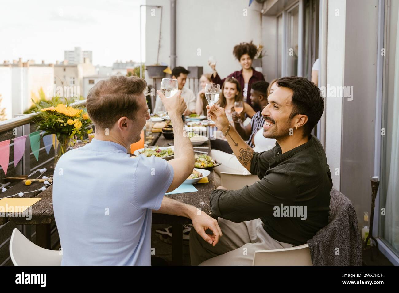 Glückliche Männer, die Getränke mit einer Gruppe von Freunden am Esstisch auf dem Balkon trinken Stockfoto