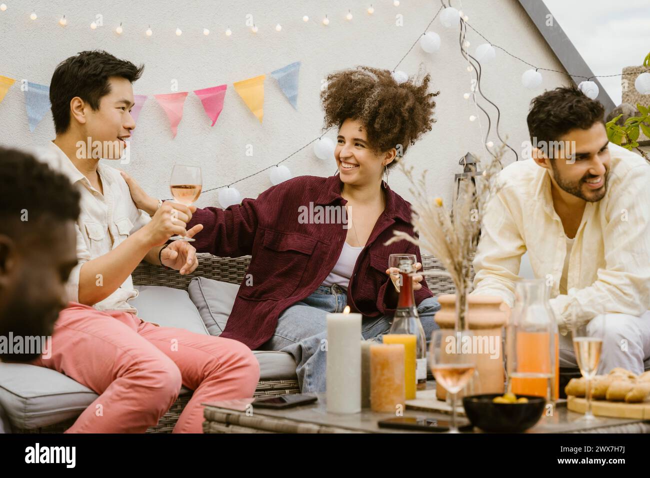 Lächelnde Frau mit Hand auf der Schulter eines männlichen Freundes, der bei einer Party auf dem Balkon ein Glas hält Stockfoto