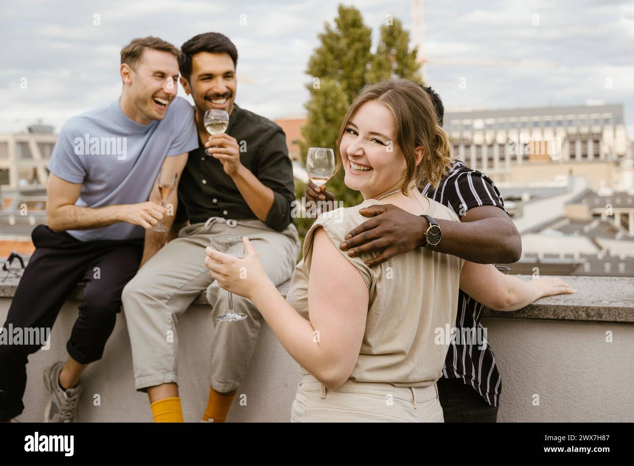 Porträt einer lächelnden Frau, die über die Schulter schaut, während sie mit männlichen Freunden auf der Dachterrasse Drinks genießt Stockfoto