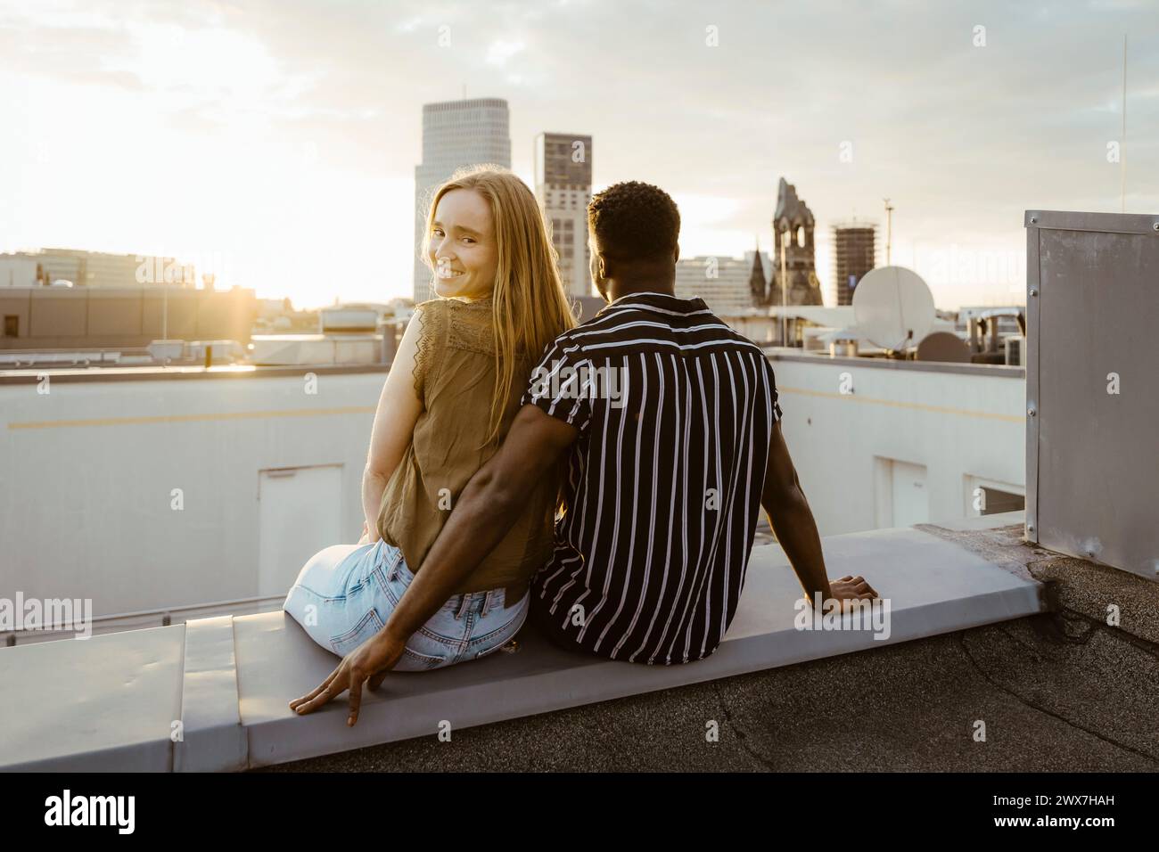 Porträt einer lächelnden Frau, die über die Schulter schaut, während sie mit ihrem Freund auf der Gebäudeterrasse sitzt Stockfoto