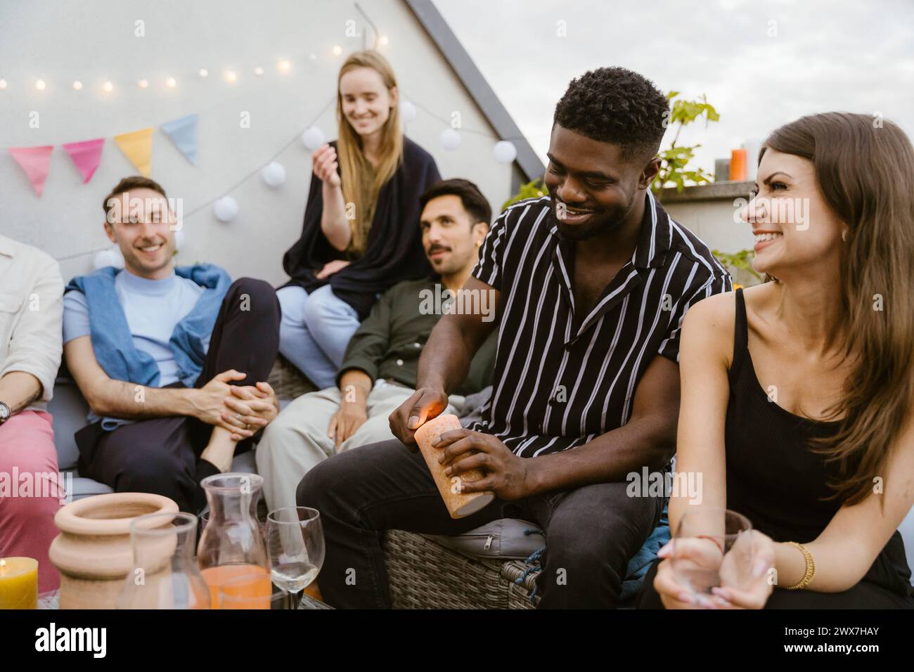 Ein lächelnder junger Mann brennt Kerze, während er mit männlichen und weiblichen Freunden auf einer Party sitzt Stockfoto