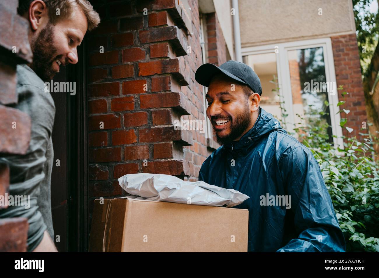 Ein lächelnder Fahrer, der mit einem Mann spricht, während er Pakete vor der Haustür zustellt Stockfoto