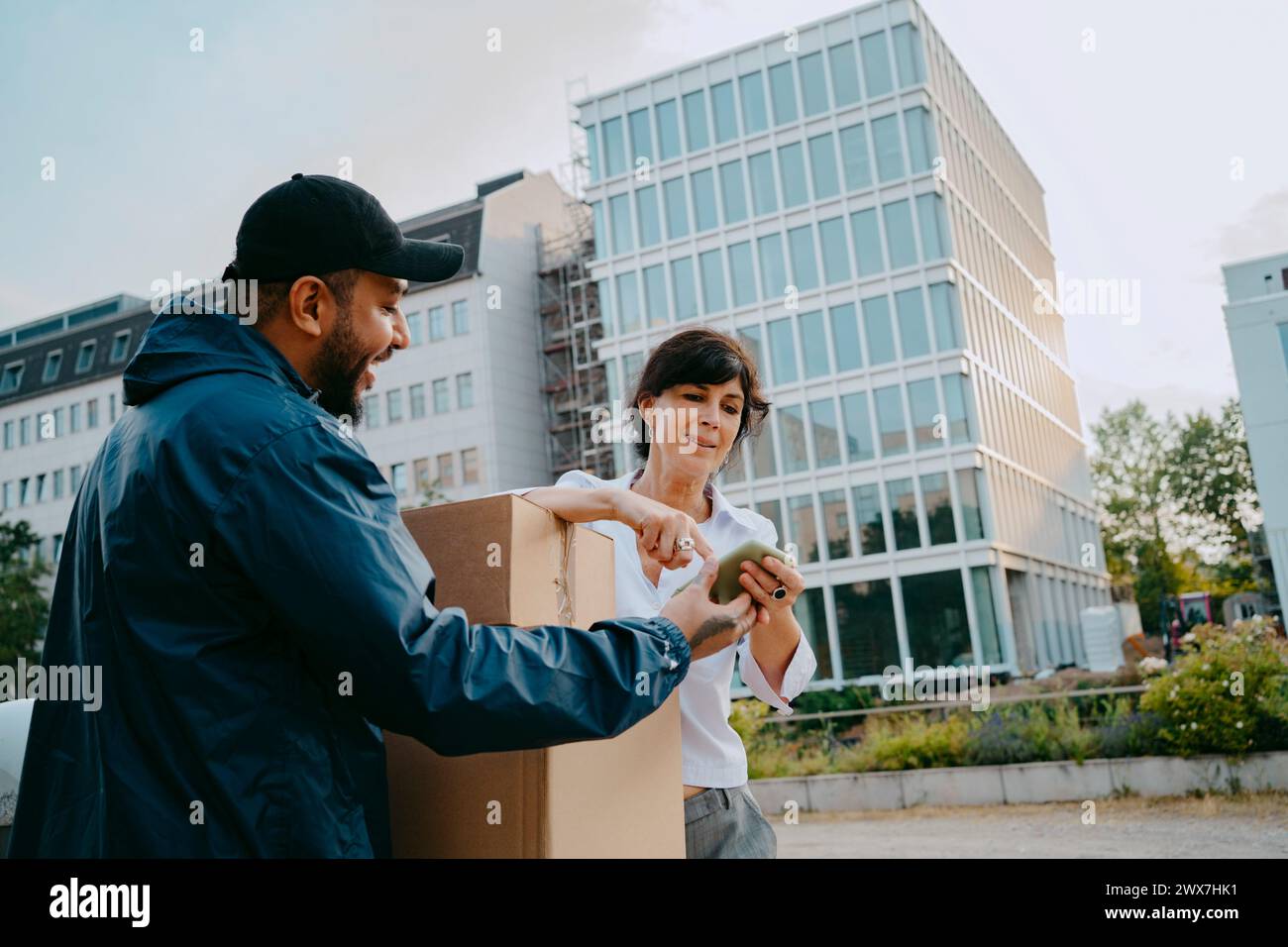 Reife Frau, die digitale Signatur macht, während sie das Paket von einer männlichen Zustellperson vor dem Gebäude erhält Stockfoto