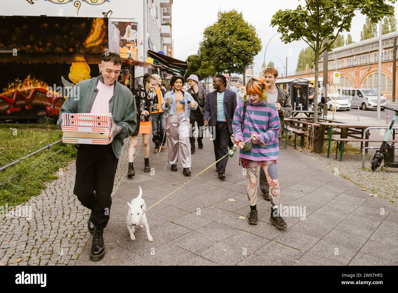 Gruppe von nicht-binären Freunden, die zusammen auf dem Bürgersteig mit Bullterrier Hund in der Stadt laufen Stockfoto
