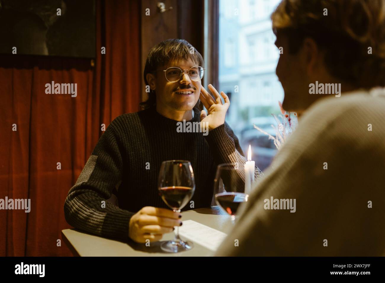 Ein lächelnder schwuler Mann hält ein Weinglas und spricht mit seinem Freund, während er im Restaurant sitzt Stockfoto