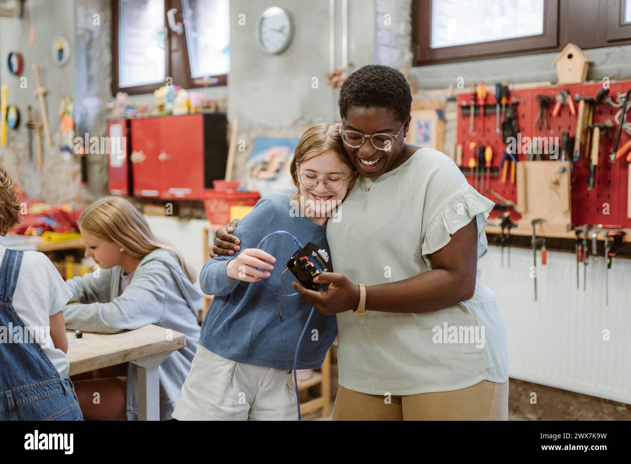 Glückliche Lehrerin, die die Schülerin bewertet, während sie in der Schulwerkstatt Elektroteile hält Stockfoto