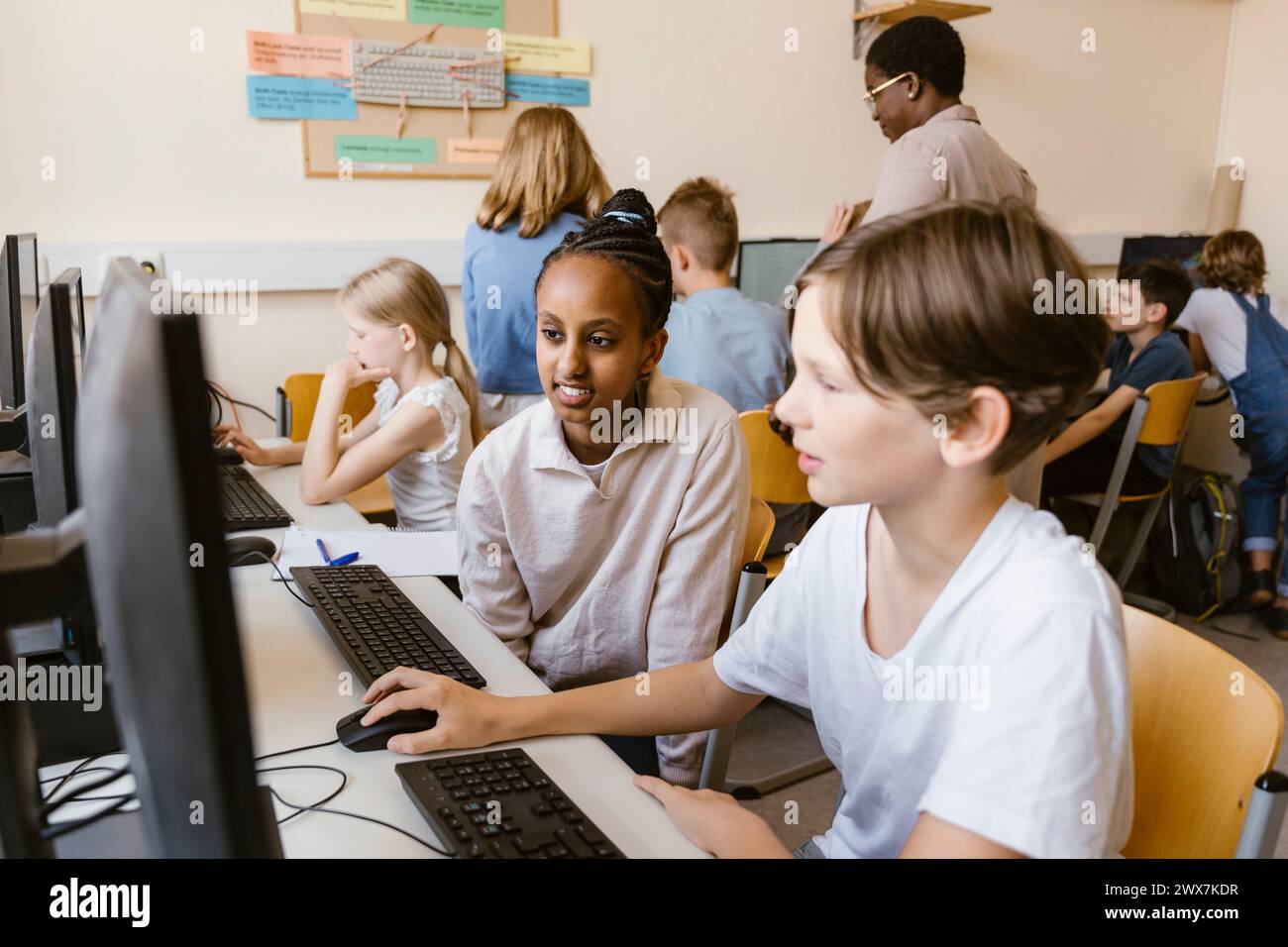 Lächelndes Mädchen, das mit einem männlichen Freund sitzt und den Computer benutzt, während er in der Schule sitzt Stockfoto