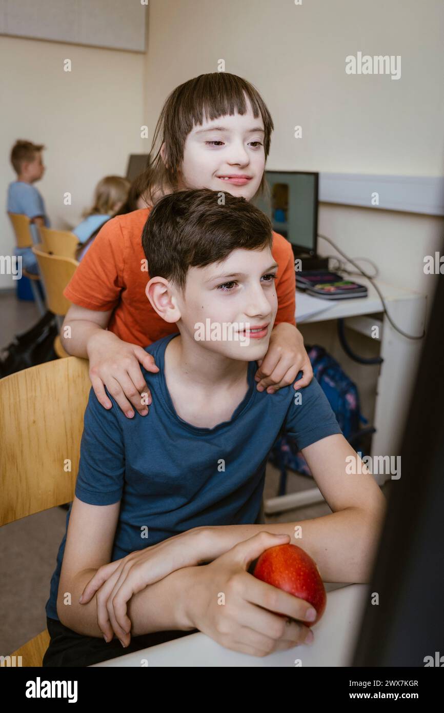 Mädchen mit Down-Syndrom in der Nähe eines männlichen Freundes, der im Computerunterricht in der Schule auf dem Stuhl sitzt Stockfoto