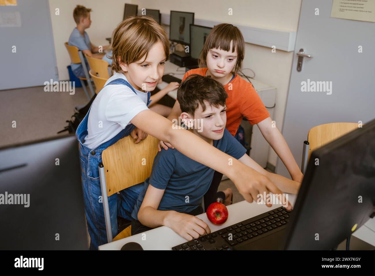 Neugieriges Mädchen, das auf den Computer zeigt, in der Nähe von Jungen und weiblicher Freundin mit Behinderung im Klassenzimmer Stockfoto