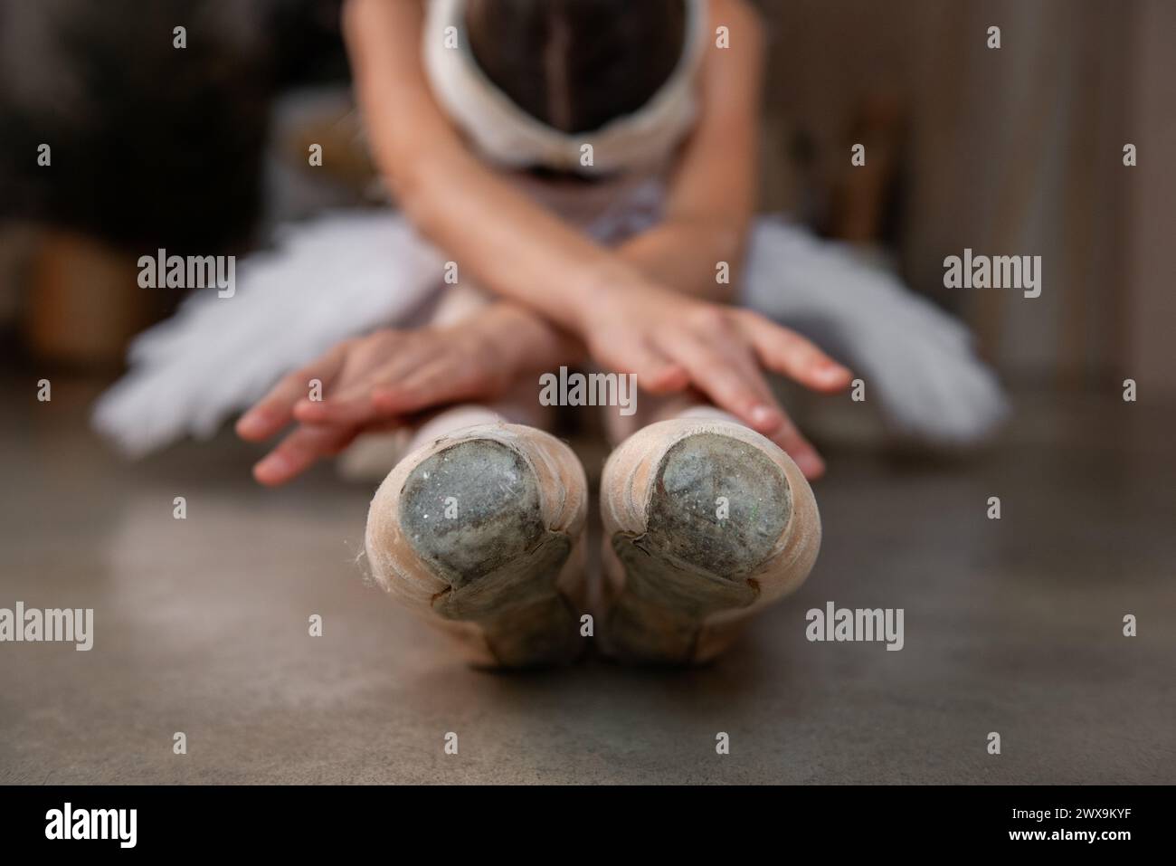 Junge Ballerina in Ballett pointe, weißes Tutu, Diadem in ruhiger Stretch, beugt sich vor, während sie auf dem Boden sitzt, im Kontrast zur weichen Kleidung. F Stockfoto