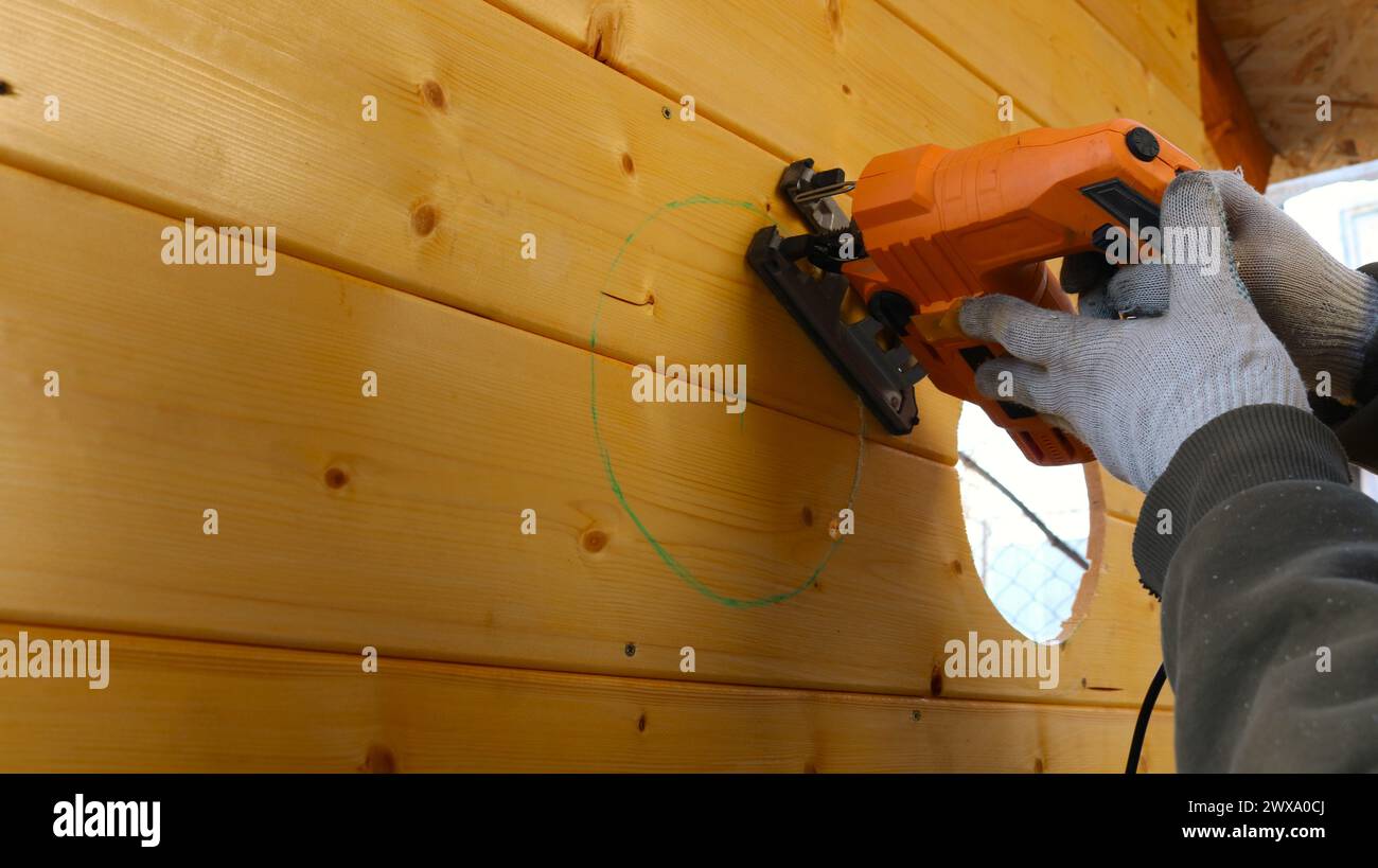 Hände in Handschuhen, die orangefarbene elektrische Ebene halten und runde Löcher auf der vertikalen Fläche der Planke schneiden, durch Kreise in der Holzwand schneiden Stockfoto