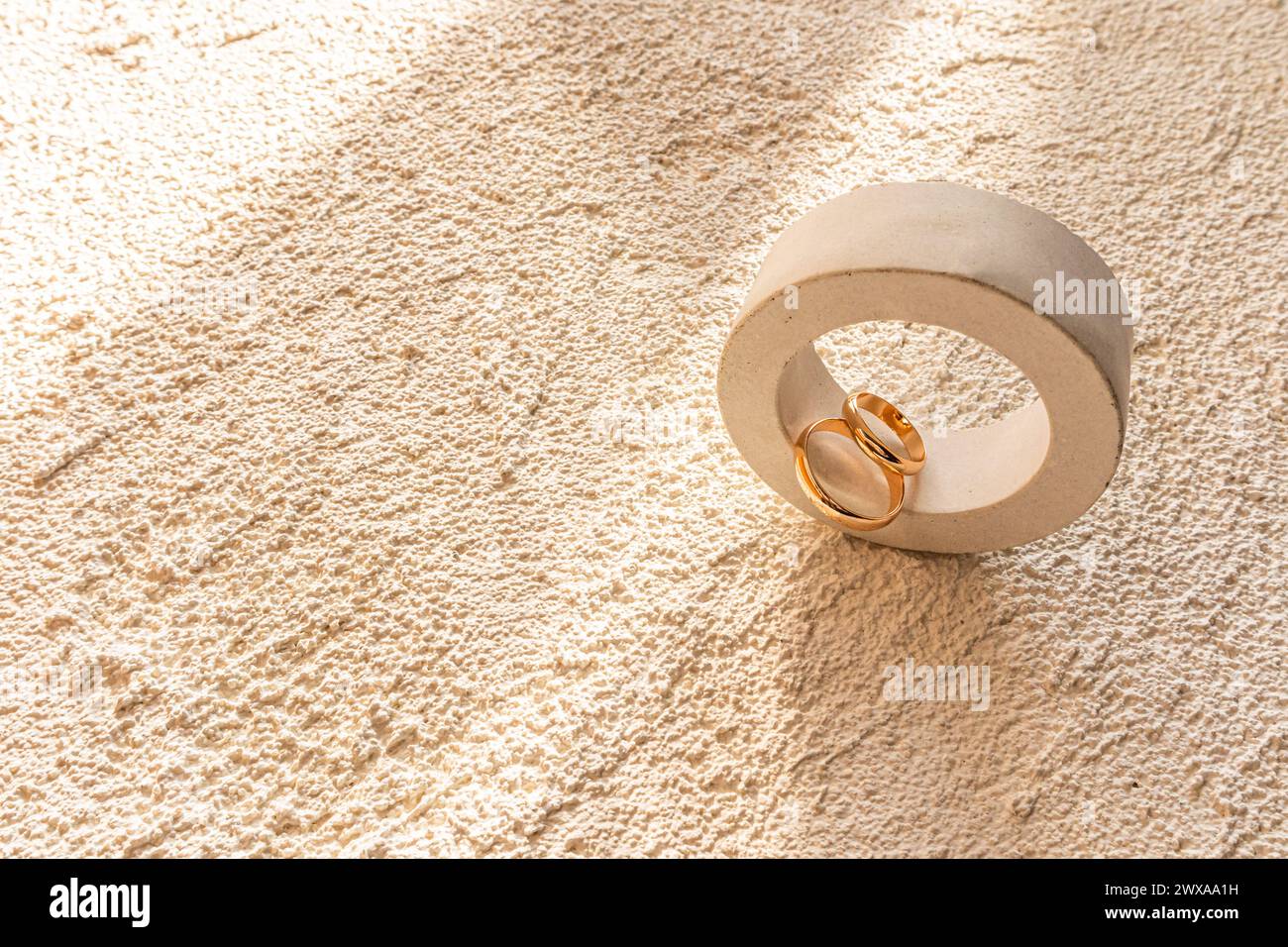 Zwei goldene Eheringe liegen in einem runden Podium aus Beton mit einem Schatten der Sonnenstrahlen. Betonreliefs Sandfarbenhintergrund. Ein Kopierbereich. Stockfoto