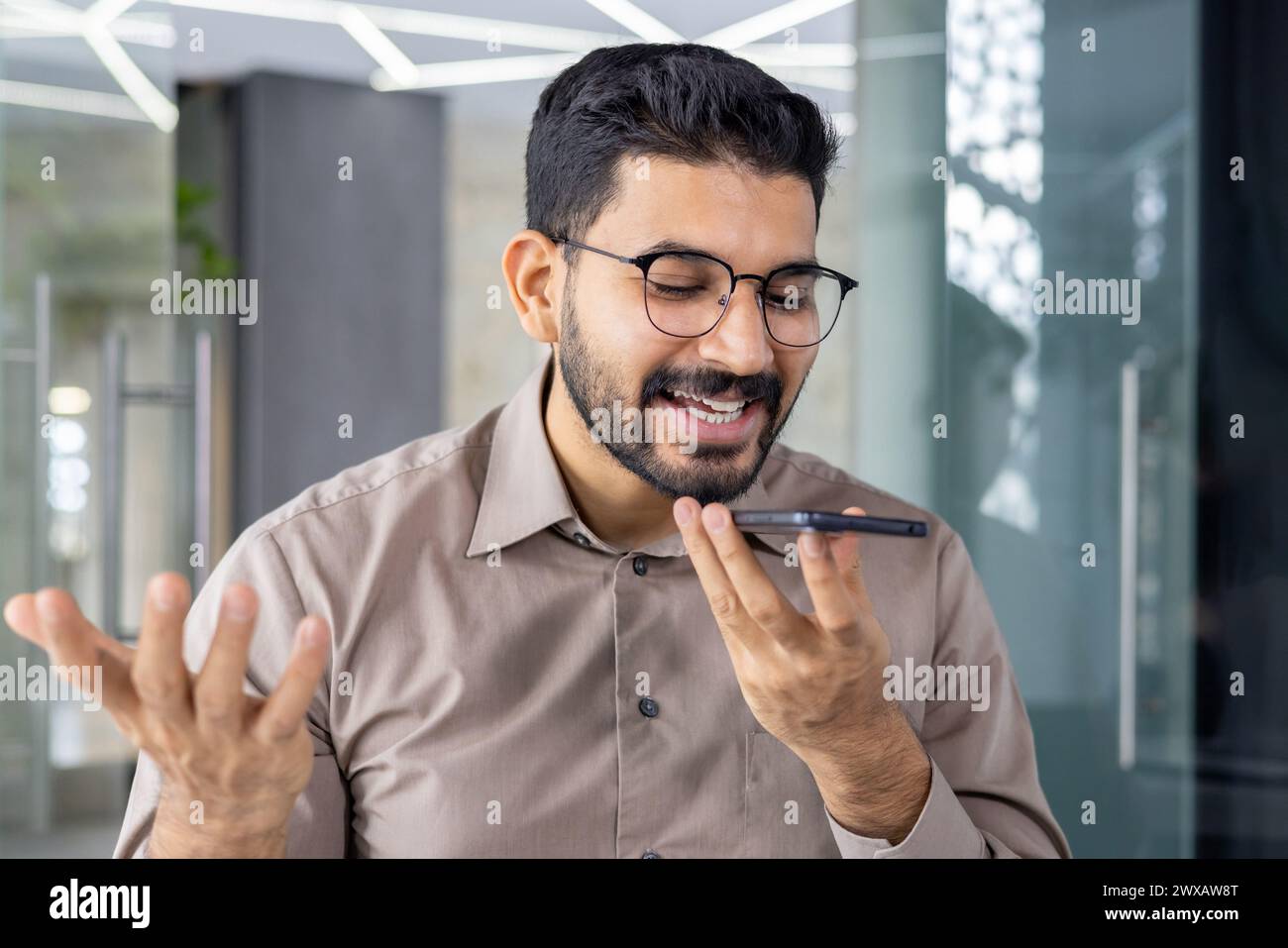Ein lächelnder junger Erwachsener, männlich, führt in einem modernen Büro ein fröhliches Gespräch mit einem freihändigen Mobiltelefon. Stockfoto