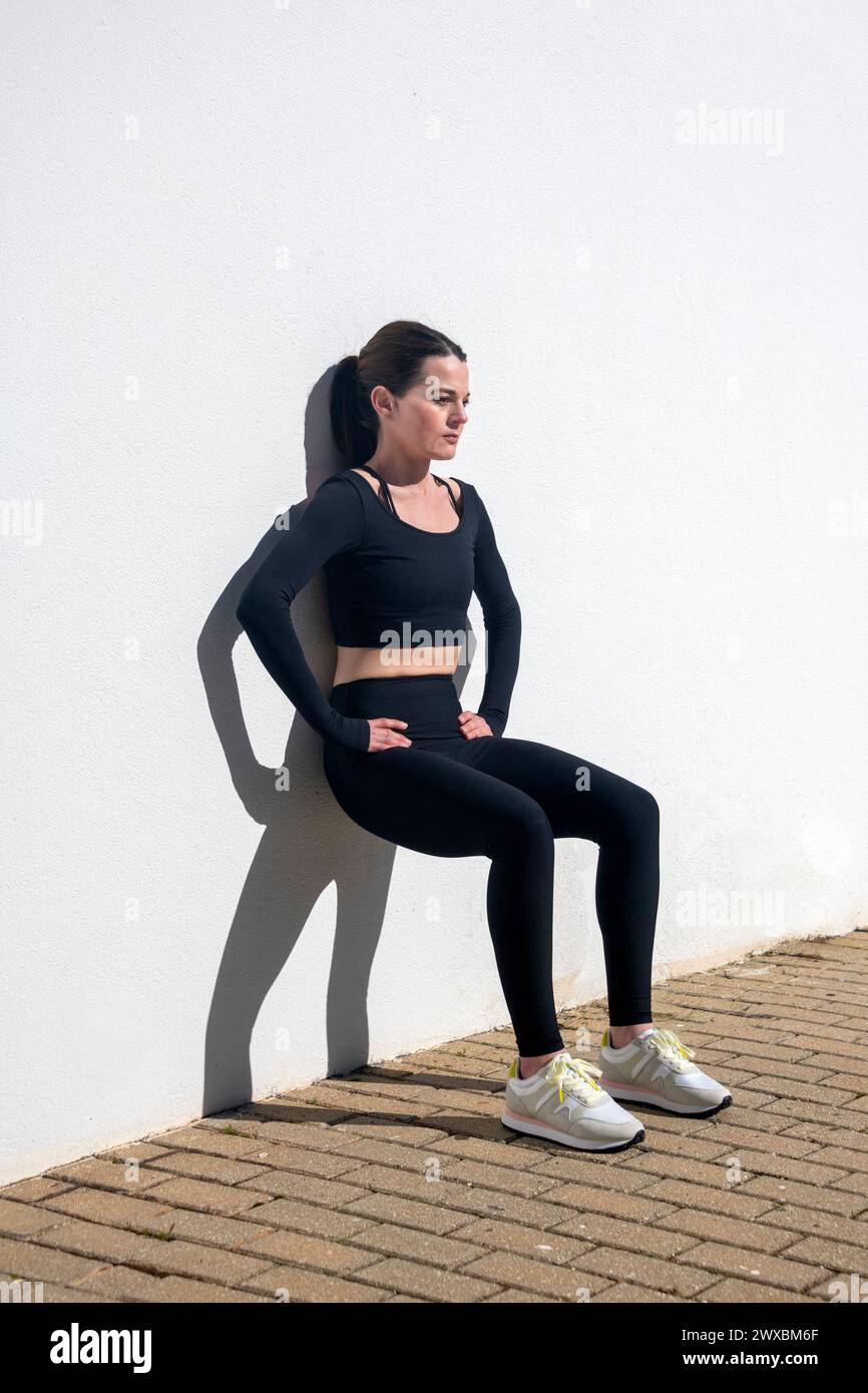 Sportliche Frau, die draußen in der Sonne Squats gegen eine weiße Wand macht. Stockfoto