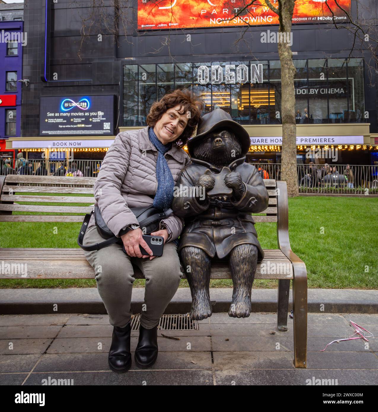 Eine weibliche Touristin, die auf einer Bank neben einer Bronzestatue des Paddington-Bären sitzt, dahinter ist ODEON Luxe eines der berühmtesten Kinos der Welt. Stockfoto