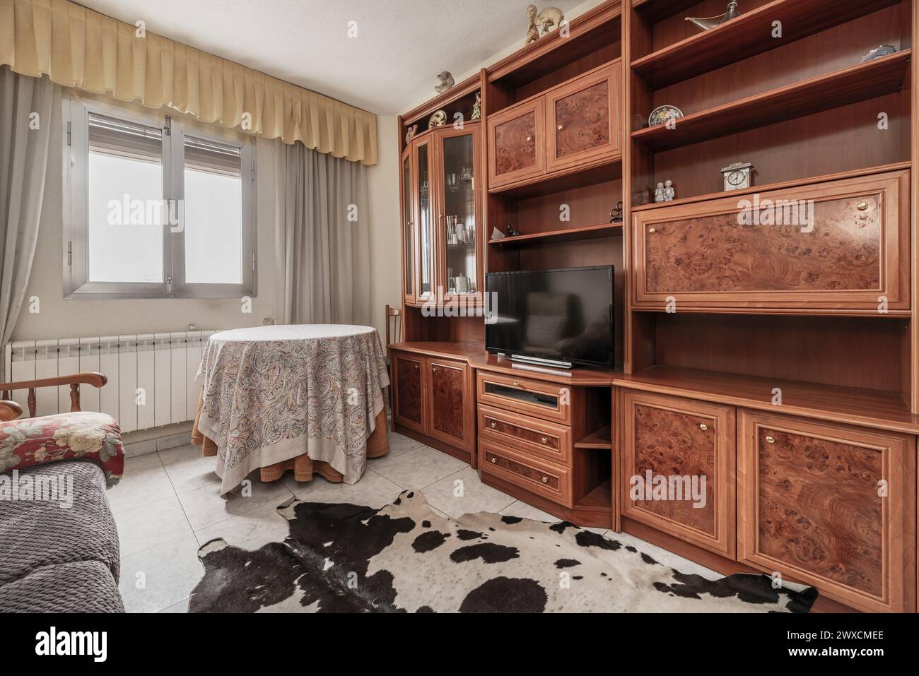 Wohnzimmer eines Hauses mit Stoffsofas in alten und schmutzigen Farben, Bücherregal aus Holzimitat, runder Liegetisch, Fenster mit Vorhängen und Boden Stockfoto