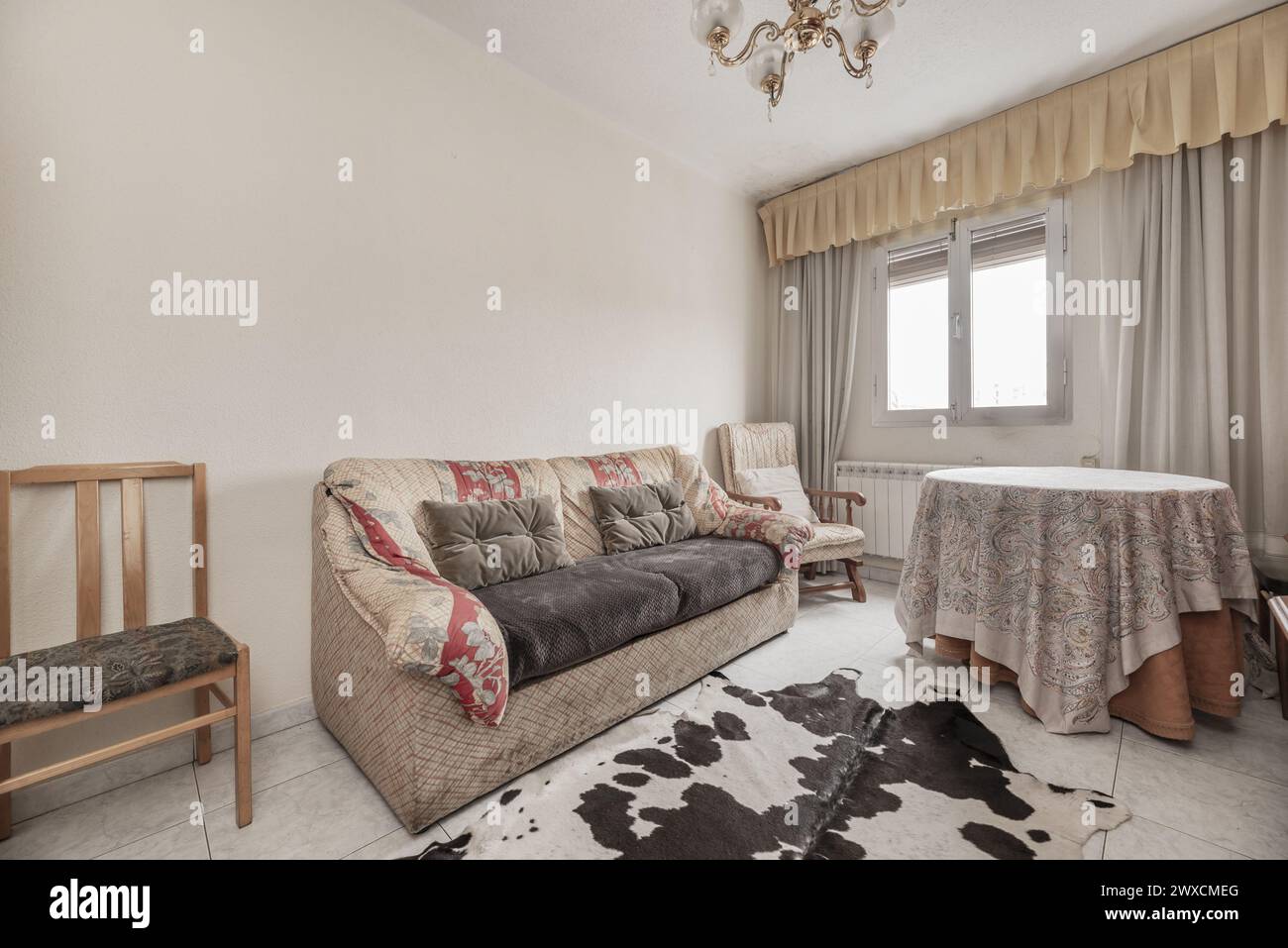 Wohnzimmer eines Hauses mit Stoffsofas in alten und schmutzigen Farben, runder Liegetisch, Fenster mit Vorhängen und Boden mit synthetischem Rindleder bedeckt Stockfoto