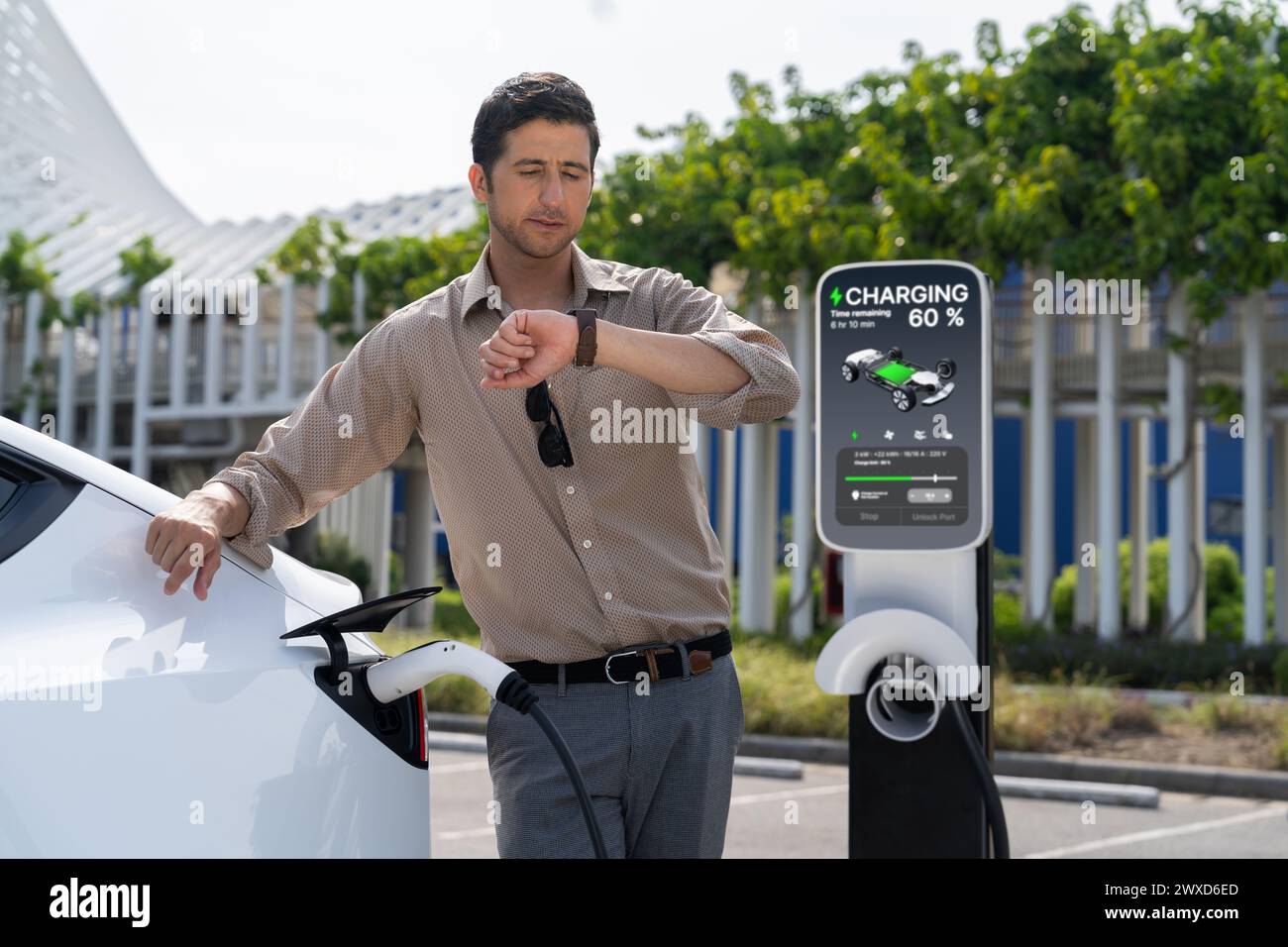 Junger Mann, der die Zeit auf Smartwatch überprüft, während EV-Ladegerät zum Aufladen der Batterie von der Ladestation auf dem Parkplatz des Einkaufszentrums in der Stadt. Aufladbares Elektrofahrzeug für Stockfoto