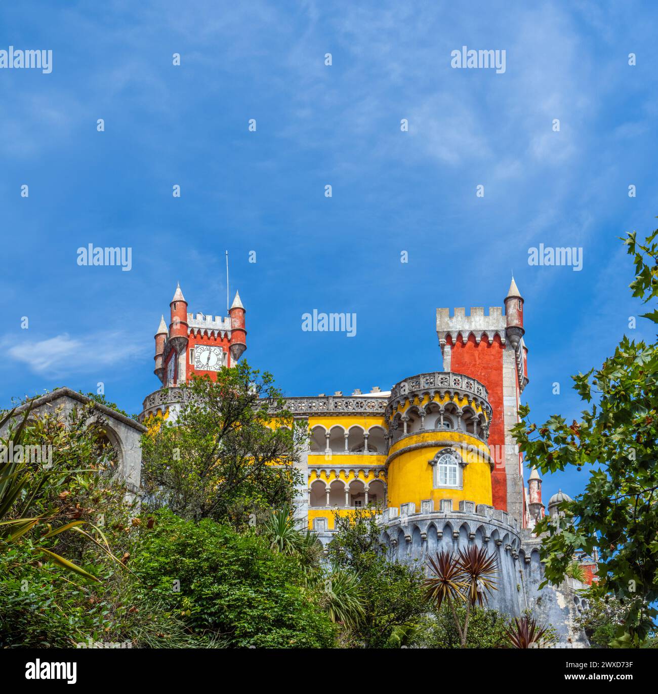 Blick von unten auf den Pena-Palast in einem bunten gelb-roten Stil auf befestigte Mauern, die mit Grün und Palmen bedeckt sind, unter einem sonnigen blauen Himmel. Stockfoto