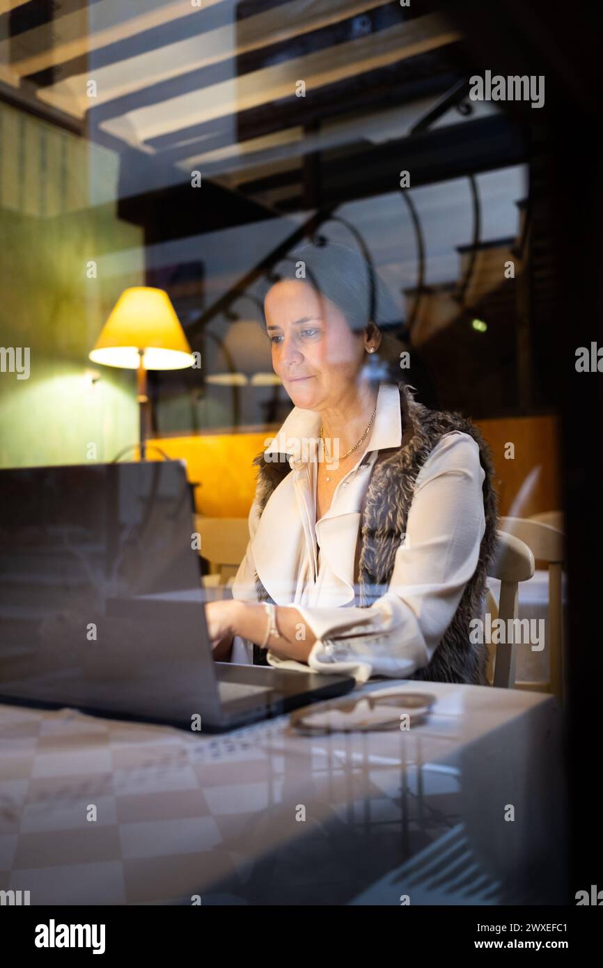 Elegante und schöne Frau mittleren Alters telearbeitet mit einem Laptop in einem Café. Sie trägt eine große, kreative Brille und das Licht aus der La Stockfoto