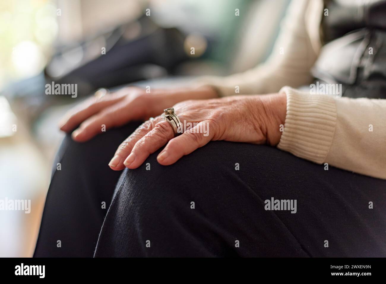 Eine ältere Frau sitzt auf einer Couch, die Hände liegen auf den Knien und trägt einen Ring auf der linken Hand. Einsamkeit und altes Randkonzept Stockfoto