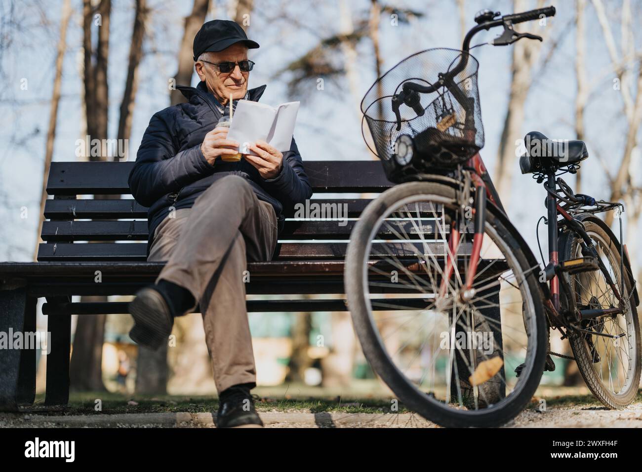 Ein älterer Herr lächelte, während er ein digitales Tablet auf einer Bank im Park benutzte, während sein Fahrrad neben ihm geparkt war und das aktive Altern darstellte. Stockfoto