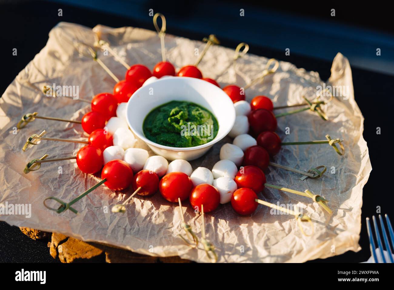 Ein herrliches Arrangement von Caprese-Spießen mit Kirschtomaten, Mozzarella-Kugeln und einem lebendigen Basilikum-Pesto-Dip auf Naturpapier. Stockfoto