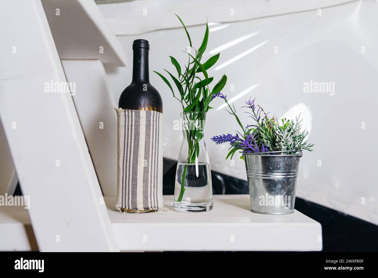 Eine einfache Einrichtung mit einer Glasvase und frischen grünen Pflanzen neben einer überdachten Weinflasche auf einer weißen Treppe. Stockfoto