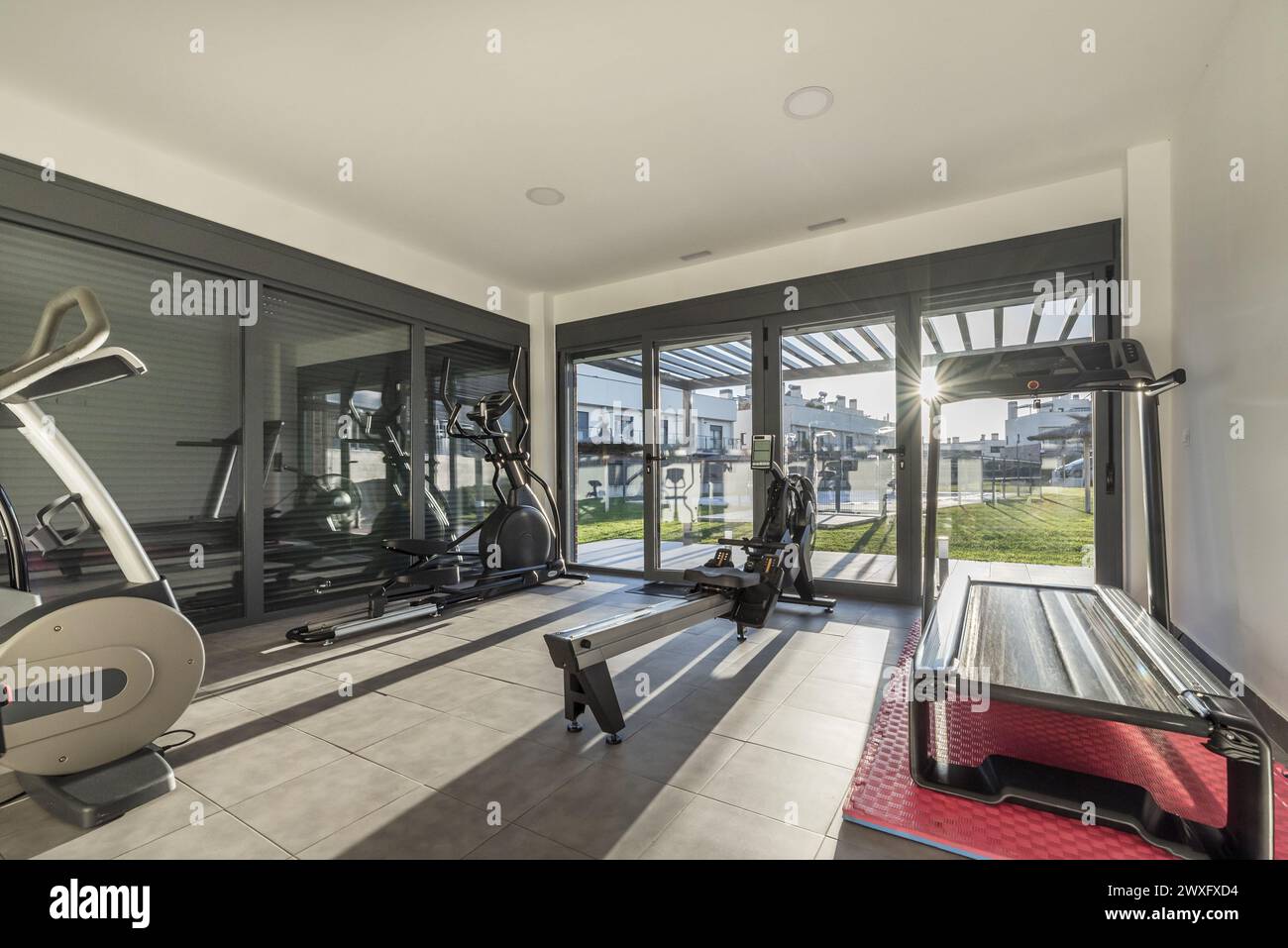 Kleiner Fitnessraum mit mehreren Fitnessgeräten in einem Gebäude mit großen Fenstern mit Fensterläden und Blick auf den Pool mit Garten. Stockfoto