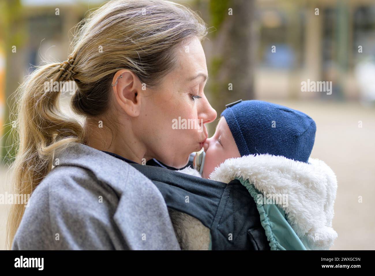 SideView einer attraktiven blonden Mutter in ihren 40ern, die ihr neugeborenes Baby liebevoll in einer Babytrage hält und ihm sanft auf die Nase küsst Stockfoto