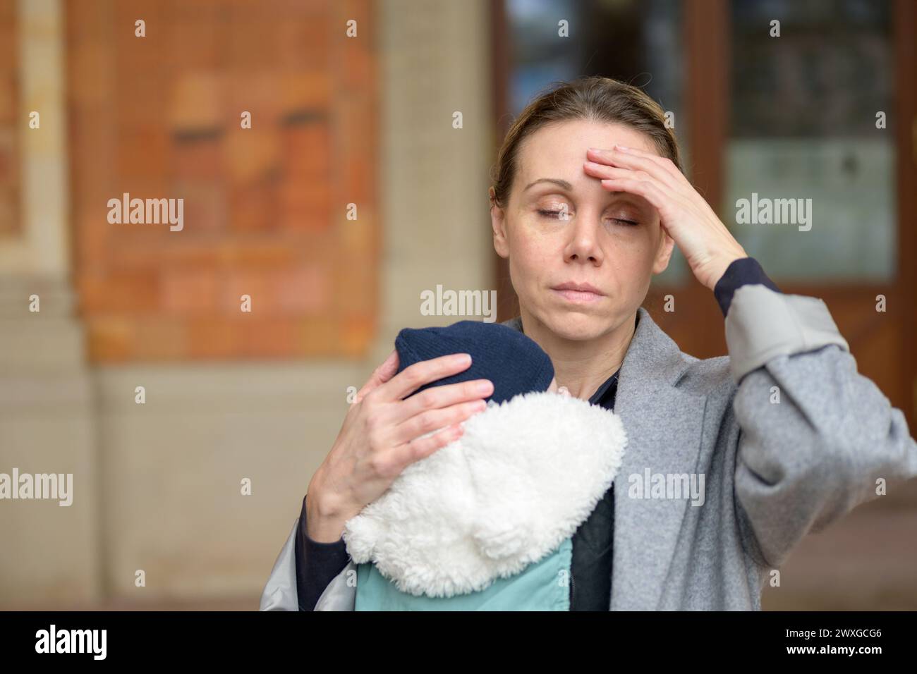 Erschöpfte und gestresste spätgeborene Mutter in ihren 40ern hielt ihr neugeborenes Baby in einer Babytrage und sah mit geschlossenen Augen genervt auf die Kamera Outdo aus Stockfoto