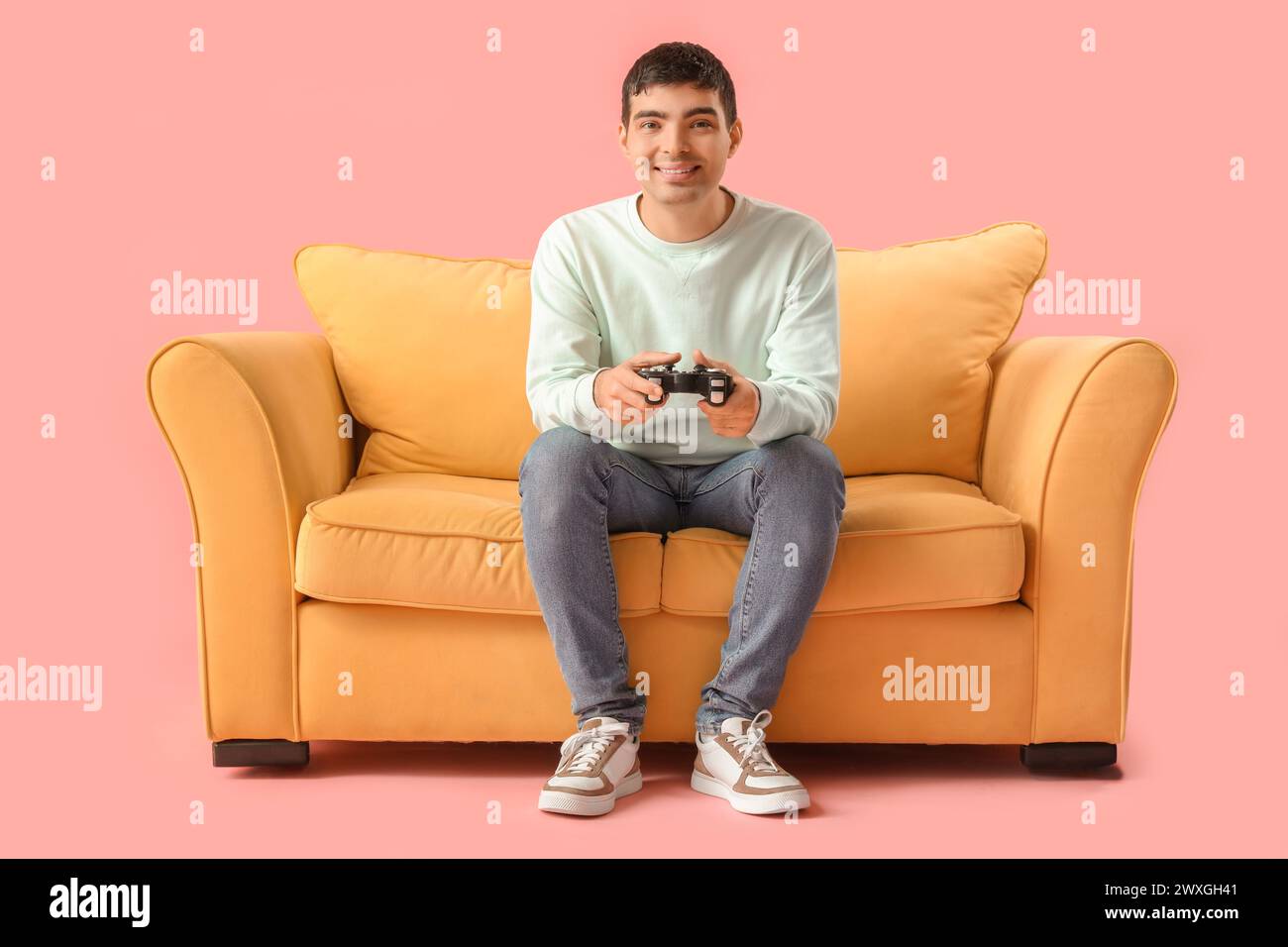 Schöner junger Mann, der Videospiel auf gelbem Sofa vor rosa Hintergrund spielt Stockfoto