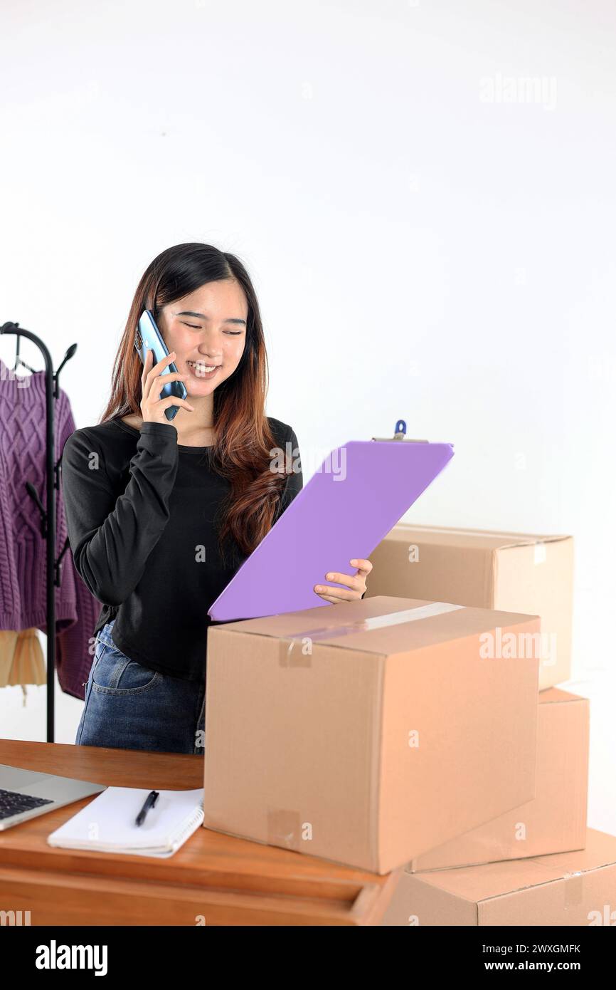 Asiatische weibliche Kleinunternehmer-Eigentümerin, Happy Face beim Anruf mit dem Smartphone und beim Überprüfen des Bestands, bei der Inventaroptimierung für Modeprodukte Stockfoto