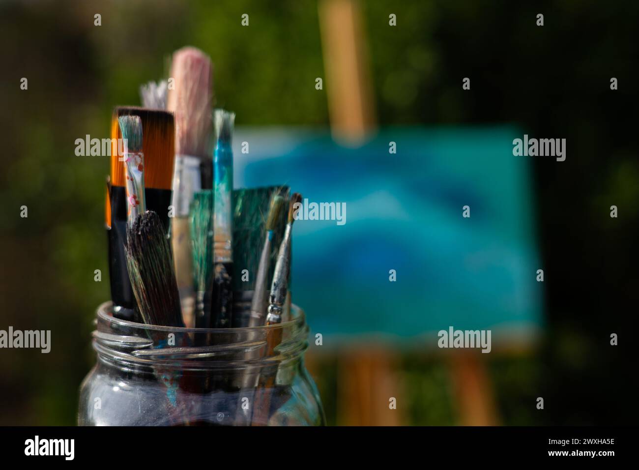 Viele Pinsel in einem Glasgefäß mit einer Staffelei und einer bemalten Leinwand dahinter Stockfoto