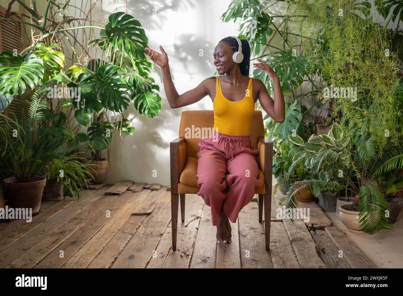Fröhliche schwarze Frau, die auf einem Stuhl sitzt und Lieblingsmusik hört, umgeben von exotischen Zimmerpflanzen. Stockfoto