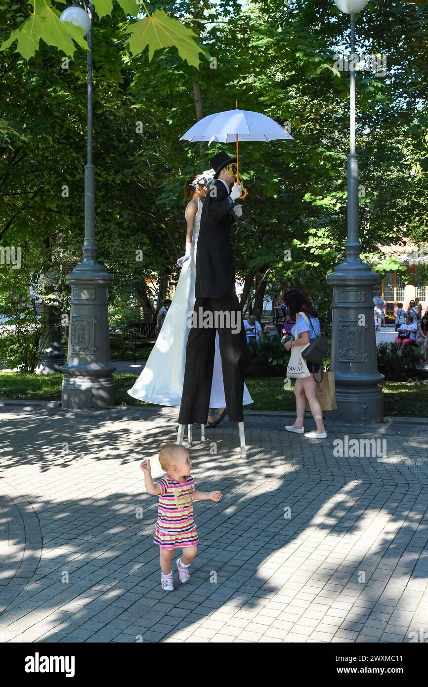 Moskau, Russland - 29. Juli 2018: Eine moderne Mutter spaziert mit ihrem Baby in einem öffentlichen Park. Ein Baby läuft überraschend in der Nähe von zwei großen Künstlern auf Stelzen in fa Stockfoto
