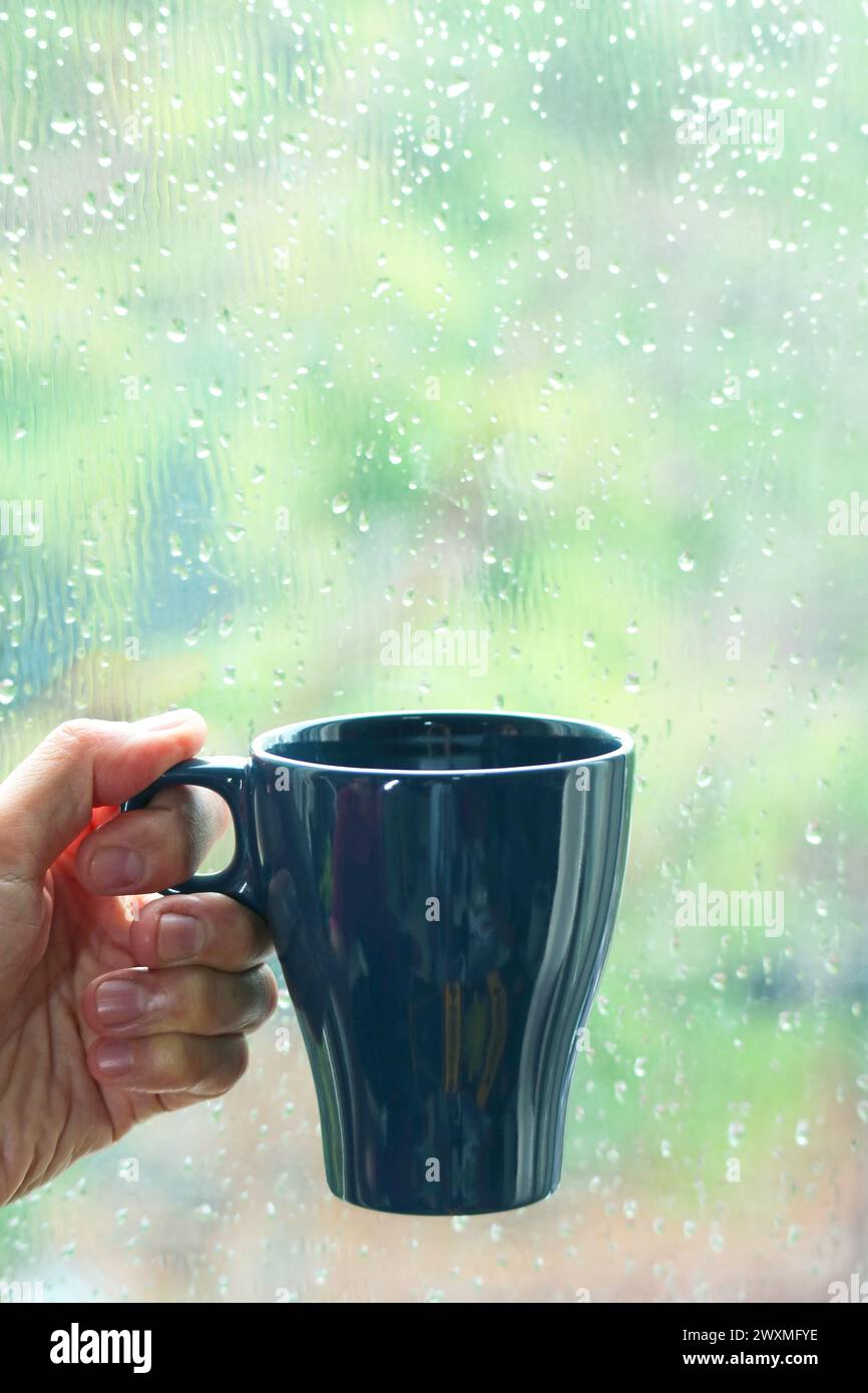 Hand hält eine Tasse Kaffee mit Fensterglas im Regen im Hintergrund Stockfoto