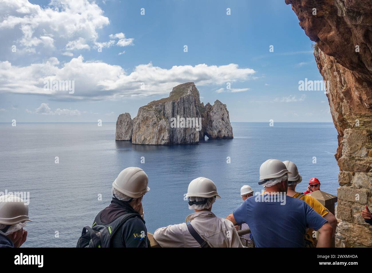 Gruppe von Touristen mit Helmen, die auf eine ferne Felsformation (Sugarloaf Island) im Meer von Porto Flavia, einer Bergbauanlage auf Sardinien, blicken Stockfoto