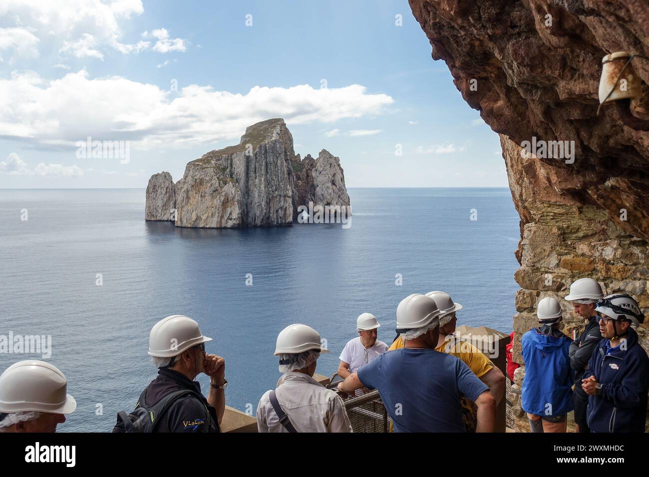 Gruppe von Touristen mit Helmen, die auf eine ferne Felsformation (Sugarloaf Island) im Meer von Porto Flavia, einer Bergbauanlage auf Sardinien, blicken Stockfoto