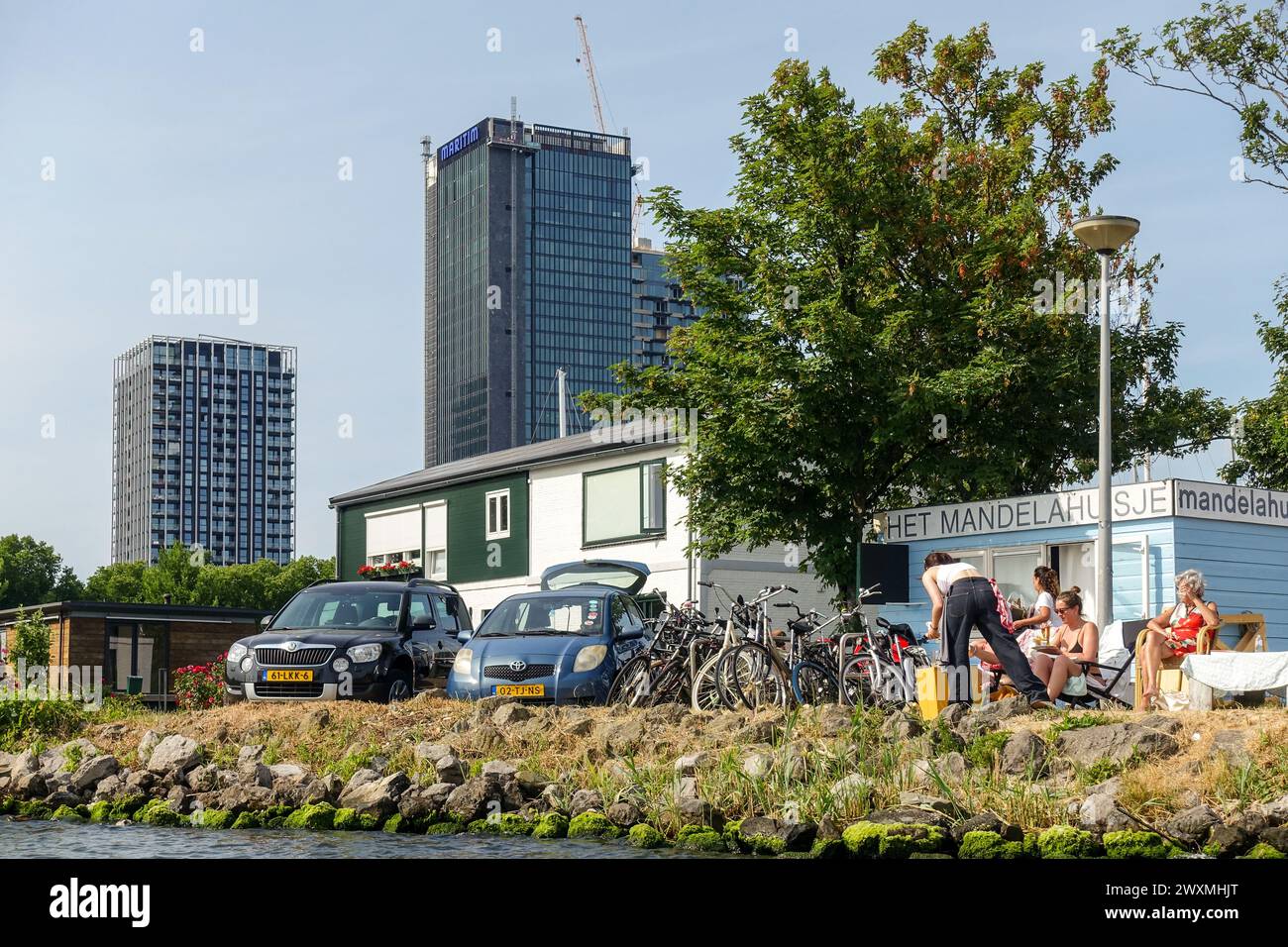 In Amsterdam Noord entspannen sich die Menschen am Wasser in der Nähe von geparkten Fahrrädern und Autos, mit modernen Hochhäusern im Hintergrund an einem sonnigen Tag Stockfoto