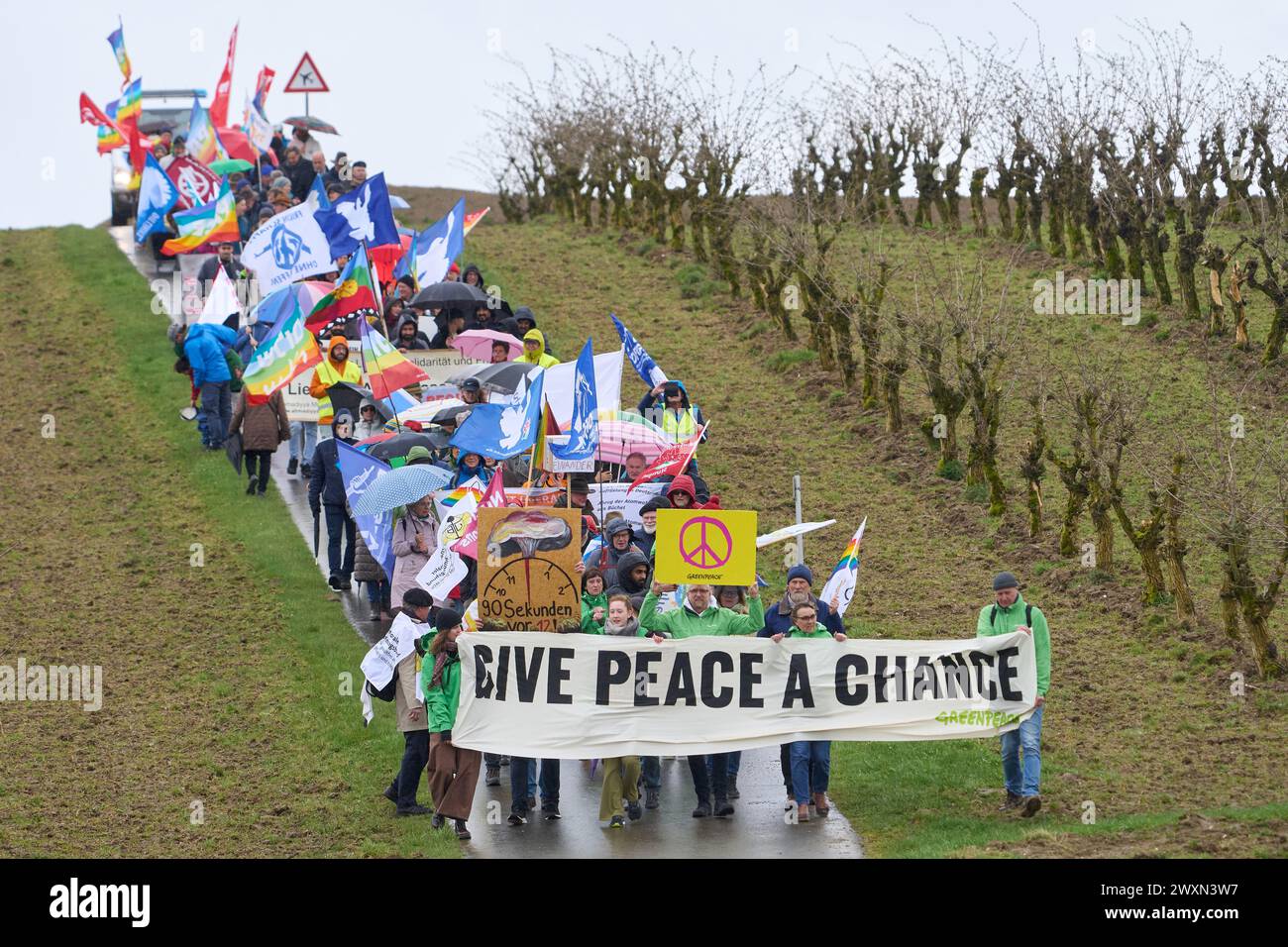 01. April 2024, Rheinland-Pfalz, Büchel: Teilnehmer des ostermarsches am Luftwaffenstützpunkt Büchel (Bezirk Cochem-Zell) laufen am Außenzaun des Stützpunktes entlang, wo US-Atomwaffen gelagert werden sollen. Auf dem Banner steht: "Gib dem Frieden Eine Chance". Foto: Thomas Frey/dpa Stockfoto