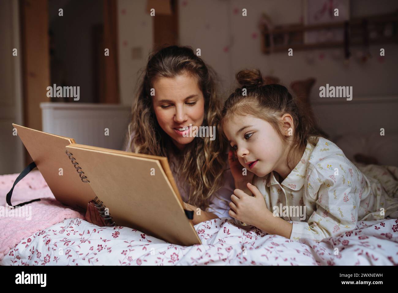 Schöne Mutter, die sich alte Familienfotos im photoalbum ansieht, mit ihrer Tochter, die im Kinderzimmer auf dem Bett liegt. Ruhiger Hygge-Familienmoment. Stockfoto