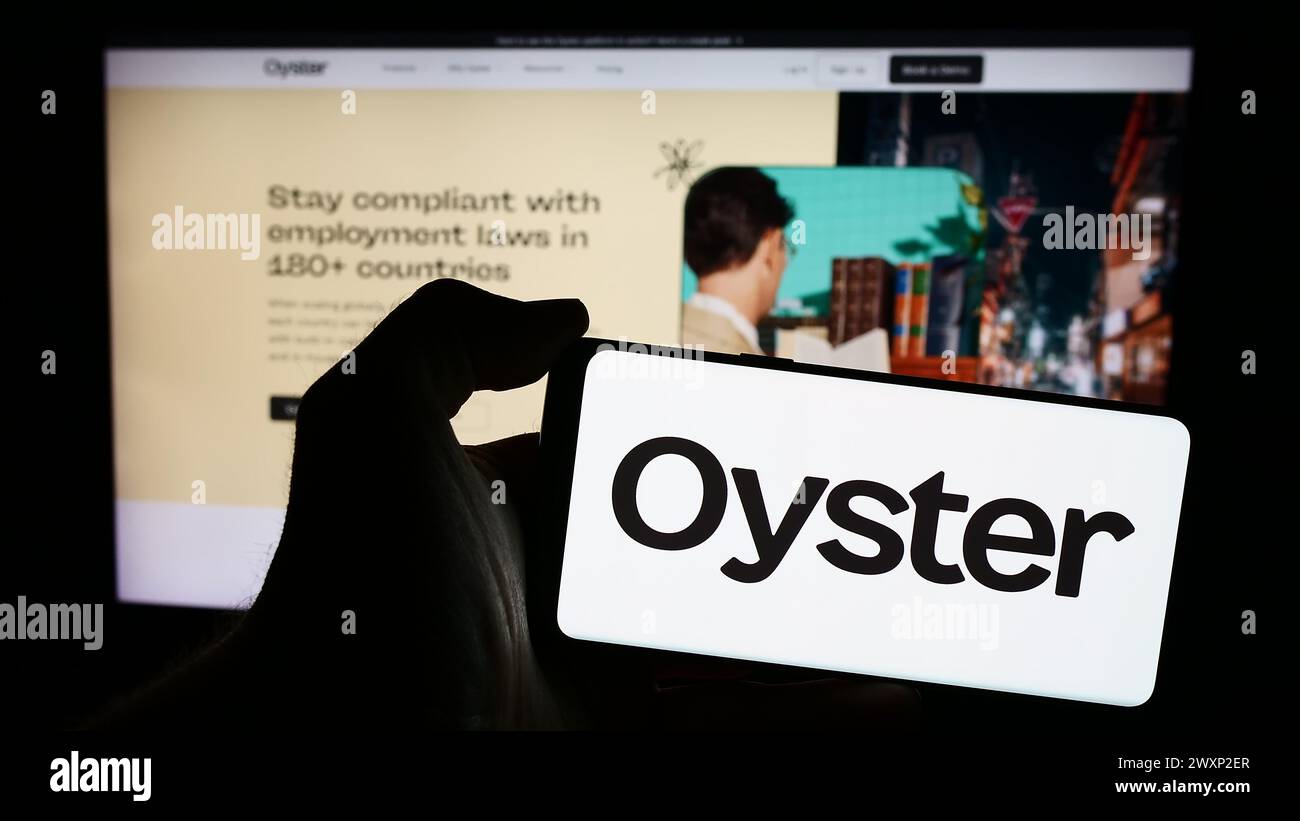 Person, die ein Mobiltelefon mit dem Logo der amerikanischen Personalplattform Oyster HR Inc. Vor der Webseite hält. Schwerpunkt auf dem Display des Telefons. Stockfoto