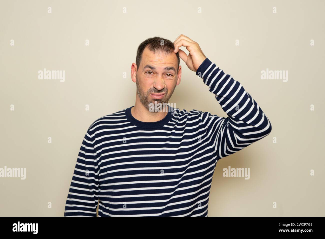 Bärtiger hispanischer Mann in seinen 40ern, der einen gestreiften Pullover trug und sich den Kopf kratzte, während er versuchte, eine Entscheidung zu treffen, Zweifel. Isoliert auf beigefarbenem Studio Stockfoto