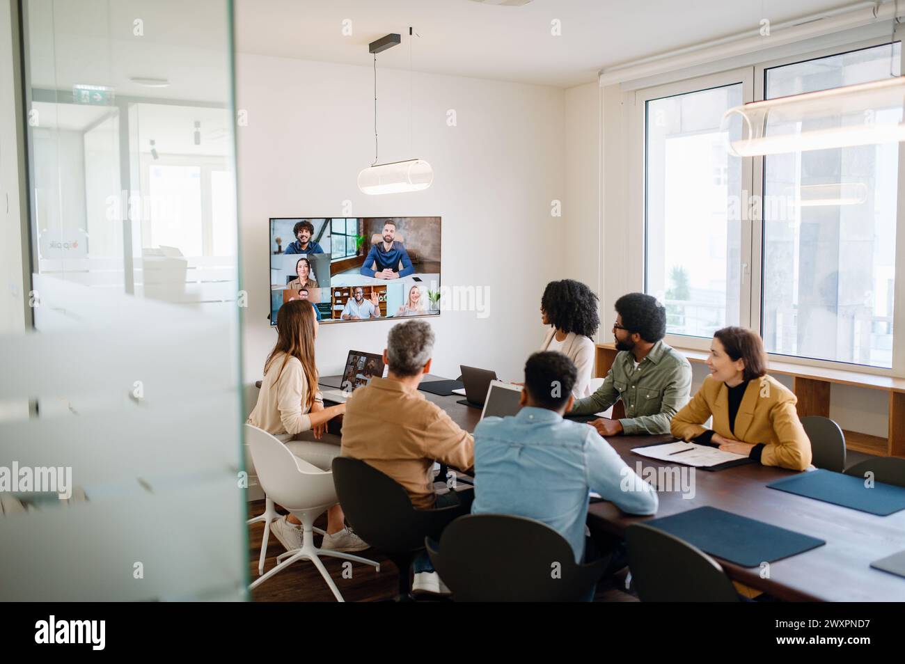 Ein Team arbeitet aktiv mit Kollegen aus der Ferne über einen großen Bildschirm zusammen und veranschaulicht das moderne hybride Arbeitsmodell, bei dem Technologie die Lücke zwischen Teammitgliedern im Büro und Remote-Mitarbeitern überbrückt Stockfoto