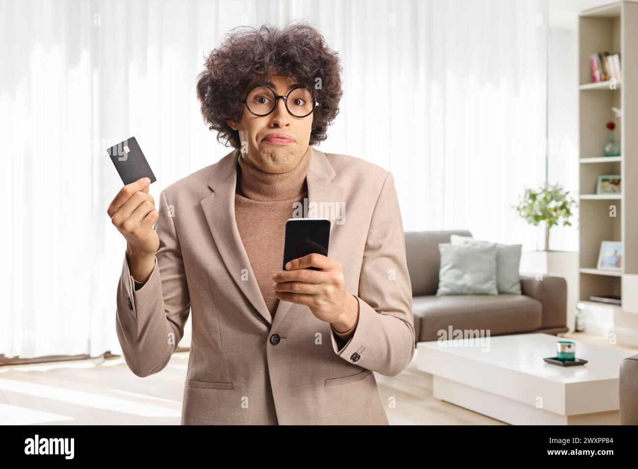 Verwirrter junger Mann, der zu Hause eine Kreditkarte und ein Handy hält Stockfoto