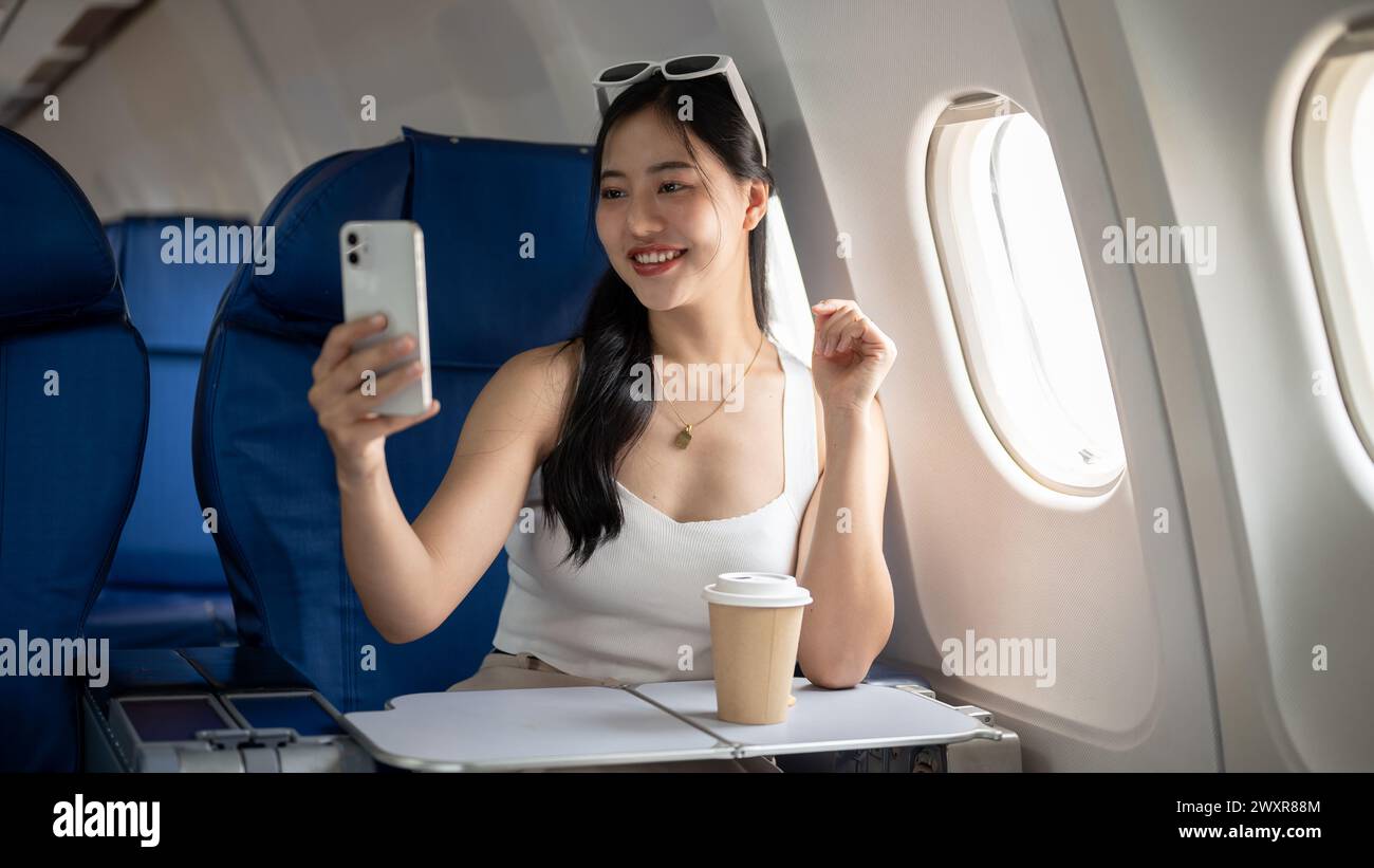Eine attraktive, fröhliche asiatische Passagierin macht während des Fluges zu ihrem Urlaub ein Selfie oder nimmt ein Video von sich selbst mit ihrem Smartphone auf Stockfoto