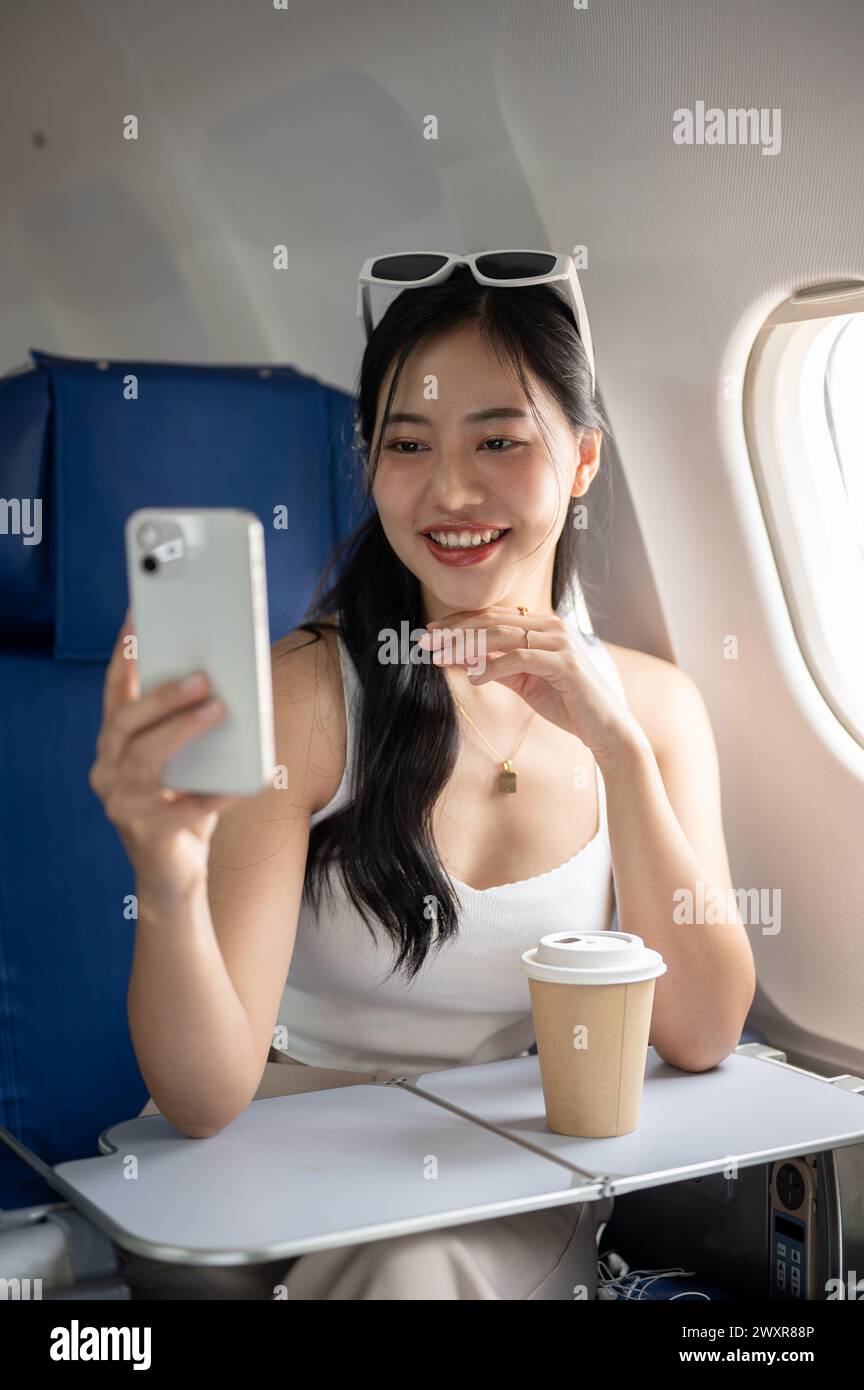 Eine attraktive, fröhliche asiatische Passagierin macht während des Fluges zu ihrem Urlaub ein Selfie oder nimmt ein Video von sich selbst mit ihrem Smartphone auf Stockfoto