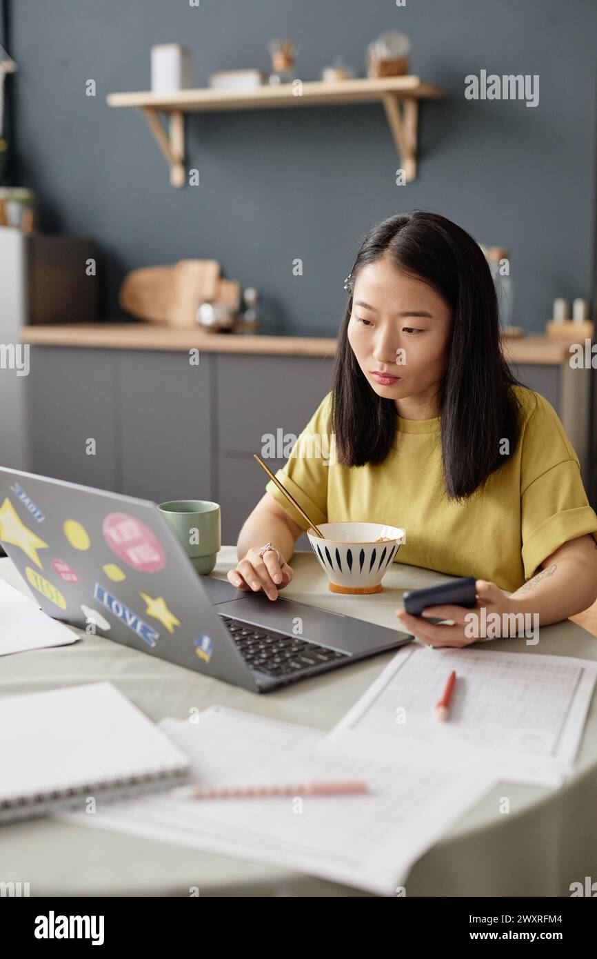 Vertikale Aufnahme einer jungen asiatischen Universitätsstudentin, die morgens beim Frühstück am Tisch sitzt und einen Laptop benutzt Stockfoto